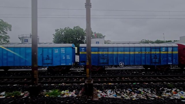 A window view from a moving train passing by stationary blue freight wagons on parallel tracks, with trees and industrial tanks in the background under a cloudy sky.