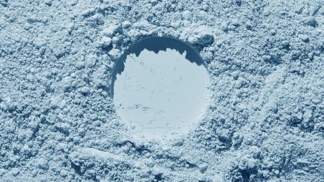 Top angle shot of pale blue natural powder surface with a circular stamp, highlighted by orbiting light that produces changing shadows