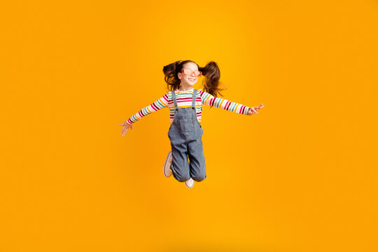 Happy girl jumping in mid-air wearing striped shirt and denim overalls on bright orange background, joyful playful childhood energy and motion