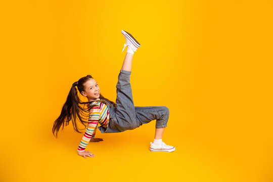Smiling girl breakdancing in denim overalls and striped shirt on bright yellow background, playful energetic kid portrait showing movement, balance and joyful youth style