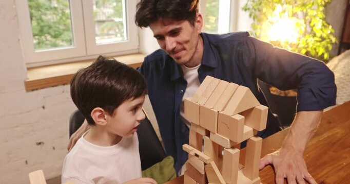 Smiling father and son completing tall wooden block tower during creative playtime. Concept of preschool education, family lifestyle media and wooden toy ads.