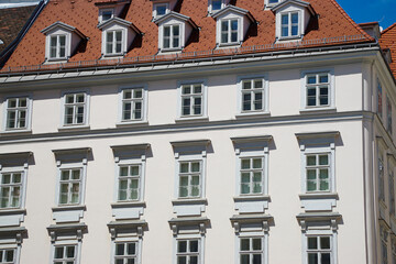 Naklejka premium Low-angle view of a historic white building with repeating windows, decorative facade details, dormer windows, and a red tile roof against a clear blue sky.