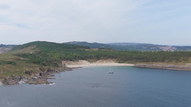 Wide aerial shot of Praia de Melide, a secluded sandy beach surrounded by lush green forest and rugged rocky coastline in Cangas, Galicia, Spain.