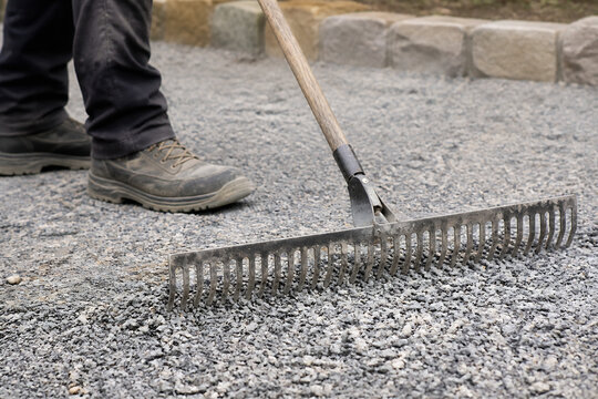 A person's feet in work boots are near a rake resting on gravel ground.
