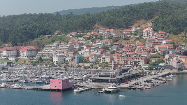 A high-angle aerial shot of the marina in Combarro, Spain, showcasing numerous docked boats and yachts, a distinctive pink waterfront building, and a hillside village with red-roofed houses.