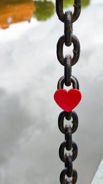 Red heart-shaped love lock on a metal chain in close-up. Romantic symbol of love and attachment against a background of water and green landscape.