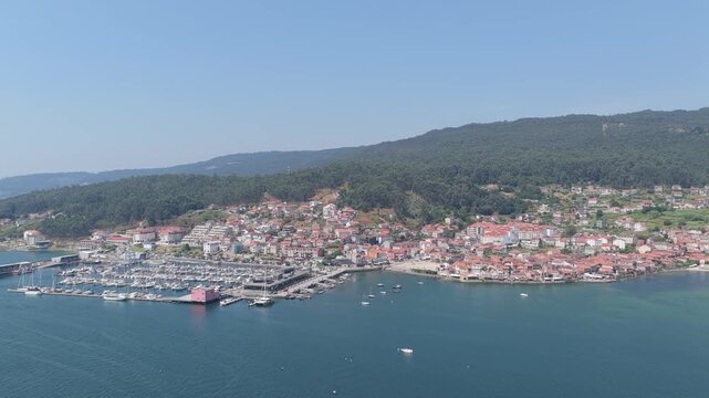 A sweeping aerial shot of the picturesque coastal village of Combarro, Spain, featuring the marina filled with boats, traditional red-roofed houses, lush green hills.