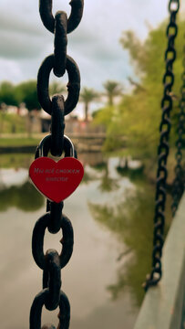 Red heart-shaped love lock on a metal chain in close-up. Romantic symbol of love and attachment against a background of water and green landscape.
