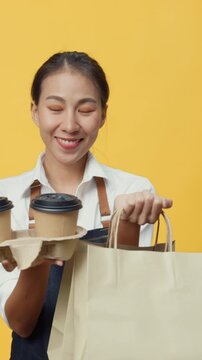Vertical Screen : Happy smile Asia waitress wear apron friendly looking at camera giving coffee or tea delicious food in craft paper bag to customer isolated on yellow background. Business takeaway.