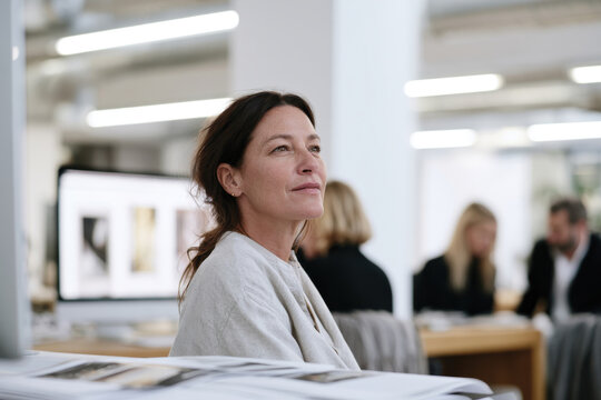 A woman in a light sweater gazes thoughtfully out of a large window in a bright, modern office space filled with design elements, embodying creativity and inspiration.