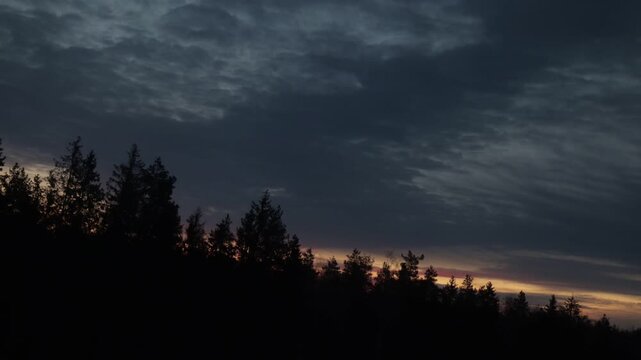 Time-lapse of sunrise over forest as dark sky becomes brighter and clouds move above tree silhouettes