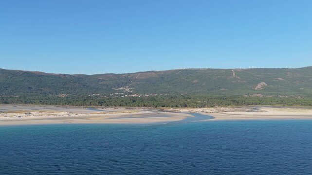 A wide aerial shot of Praia de Carnota in Carnota, Spain, showcasing the expansive sandy beach, turquoise Atlantic waters, and lush green mountains in the background under a clear blue sky.