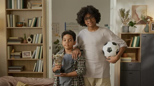 Portrait of Black child holding trophy and Black teenager holding soccer ball indoors. Video is perfect for promoting youth sports leagues, sibling support, and family achievement campaigns