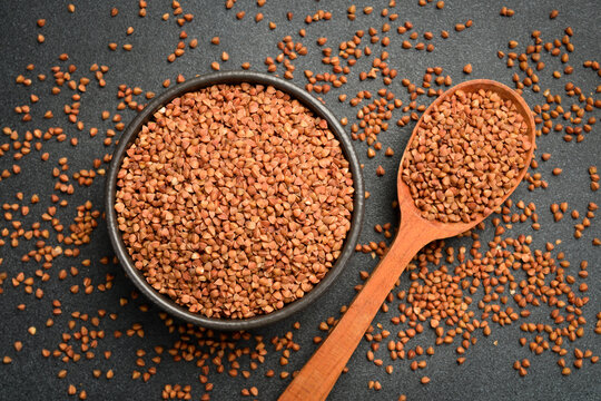Raw roasted buckwheat groats fill a round dark bowl and spoon on a stone surface. Uncooked brown grains are scattered all around. Top view horizontal photography.
