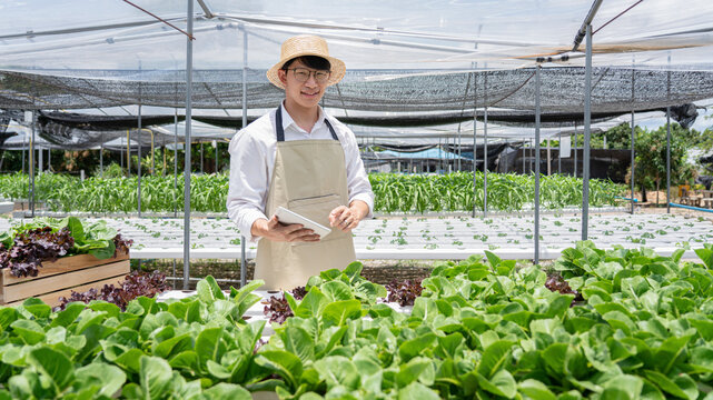 A man wearing a straw hat stands in front of a greenhouse filled with plants