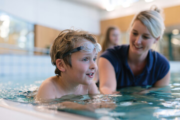 Fototapeta premium A happy child wearing swim goggles engages in swimming lessons, showcasing enthusiasm for learning in a vibrant aquatic environment under the attentive guidance of a friendly instructor.