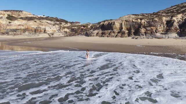 A beautiful, slow-moving drone shot gently circling around a young woman standing on the sandy shores of Praia dos Coxos, framed by the dynamic ocean waves and distant coastal cliffs