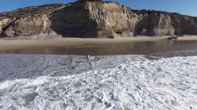 A captivating drone shot tracking and circling around a young woman as she explores the sandy beach near massive, stunning rock formations and cliffs at Praia dos Coxos, Portugal
