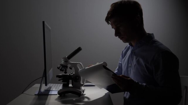 Scientist concentrating on writing research notes and analyzing data in a dimly lit laboratory, surrounded by a microscope and a computer, representing dedication to scientific discovery and study