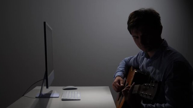 Man creating music or practicing guitar, sitting at a desk in a dimly lit room with a modern computer setup, reflecting personal time and dedication to his craft
