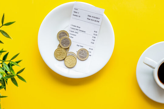 Pay restaurant bill by cash. Reciept and coins on plate on yellow background top view