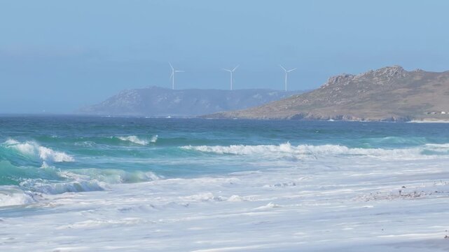 Scenic footage of Praia de Traba beach in Spain, showing turquoise waves rolling onto a sandy shore with white sea foam. In the background, rugged hills rise up, topped with modern wind turbines.