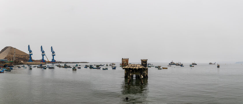 View of Puerto Chancay with small fishing boats anchored near an old wooden pier and industrial cranes in the background under an overcast sky in Chancay, Lima Region, Peru.