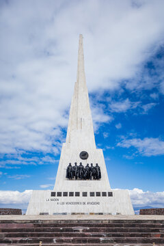 View of the Pampa De La Quinua monument featuring a tall white stone obelisk and bronze statues of soldiers against a blue sky with clouds in Quinua, Ayacucho, Peru.