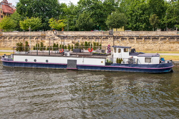 A residential houseboat, canal boat, converted barge moored alongside a stone wall, lower white cabin, rectangular windows and an upper deck railing decorated with a bright orange horseshoe life ring. © Jacek