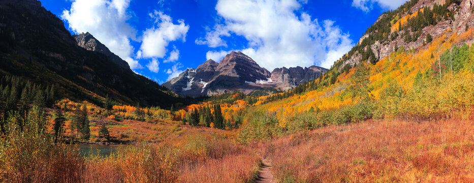 View of the Maroon Bells mountain peaks surrounded by vibrant autumn foliage and golden aspen trees under a bright blue sky in Pitkin County, Colorado, United States.