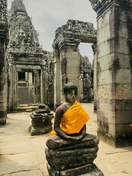 View of a Buddha statue draped in a vibrant orange robe within the ancient stone ruins of Bayon Temple, featuring intricate carvings in Krong Siem Reap, Siem Reap Province, Cambodia.