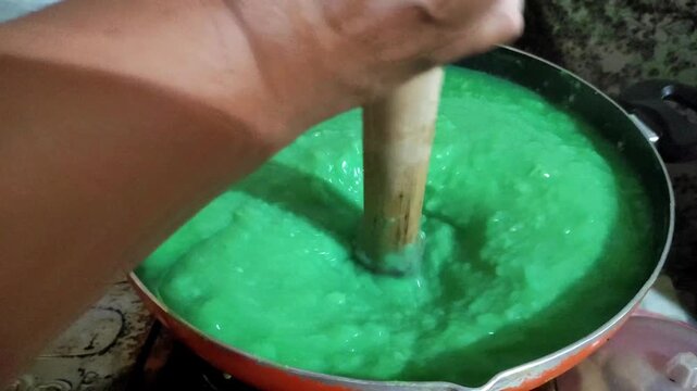 Close up of hand stirring thick vibrant green traditional pandan dessert paste in a cooking pan using a wooden pestle helper