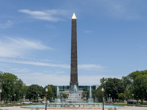 View of the Obelisk Square monument with its central fountain, surrounding green trees, and clear blue sky in Indianapolis, Indiana, United States.