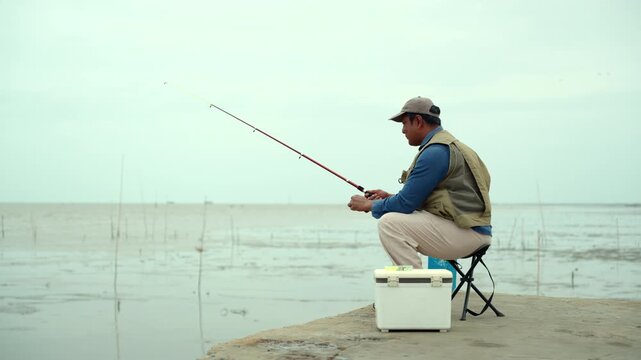Happy senior man sitting by the sea and fishing with a rod after retirement. Active aging, healthy longevity, outdoor leisure, fulfilling retirement. Life insurance and financial security concept.
