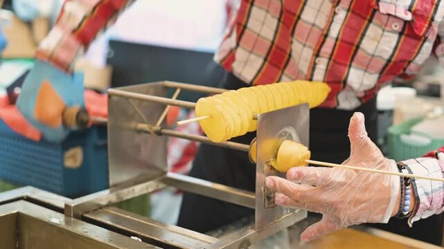 A person wearing gloves prepares a potato using a spiral cutter in a busy food market. This activity showcases the popular method of creating spiral potato snacks