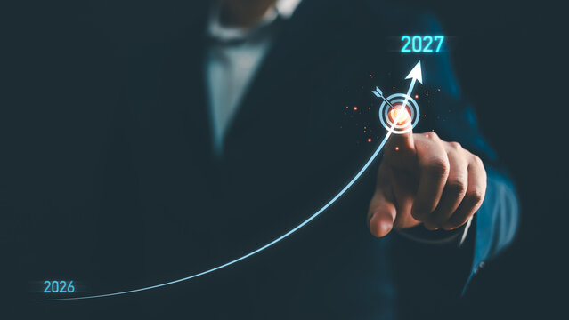 Businessman hand touching target icon with growing arrow graph from year 2026 to 2027. Concept of business goal achievement, future vision, planning strategy, success and investment growth