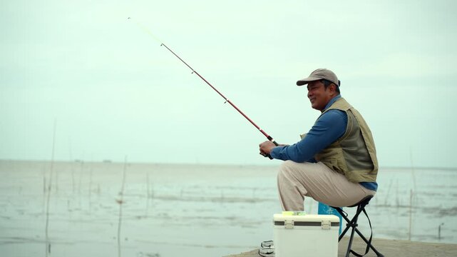 Happy senior man sitting by the sea and fishing with a rod after retirement. Active aging, healthy longevity, outdoor leisure, fulfilling retirement. Life insurance and financial security concept.
