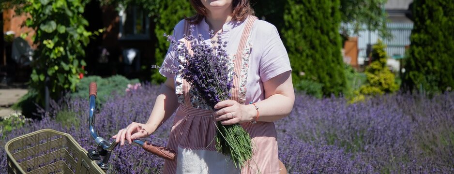 Woman in casual attire holding a lavender bouquet, standing beside a bicycle in a lush garden. Her serene expression complements the vibrant purple lavender around her