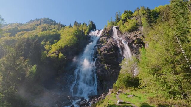 Mountain waterfall meadow trees water, gimbal stabilizer shot. Low angle view moving from left to right tracking high alpine cascade, green field and forest.