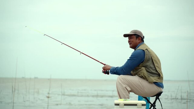 Happy senior man sitting by the sea and fishing with a rod after retirement. Active aging, healthy longevity, outdoor leisure, fulfilling retirement. Life insurance and financial security concept.
