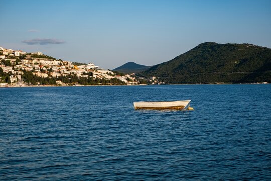 White rowboat floating on blue Adriatic water beside a coastal town and forested mountains in Neum, Bosnia and Herzegovina