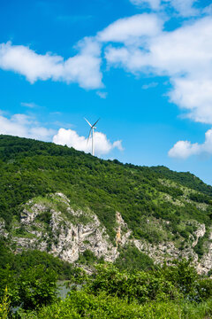 Single wind turbine rising above a green rocky mountain landscape in a rugged natural setting beneath bright blue sky and drifting white clouds