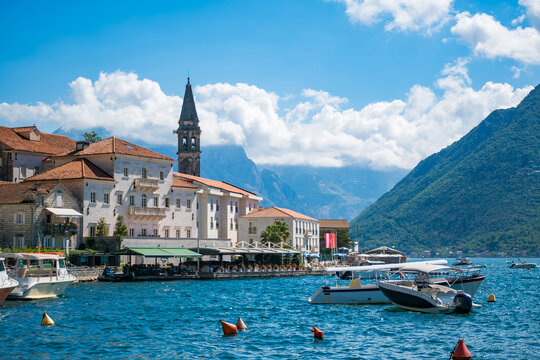 Waterfront view of Perast, Montenegro, showing a stone church bell tower, terracotta rooftops, moored boats on the blue bay, and mountains beneath clouds