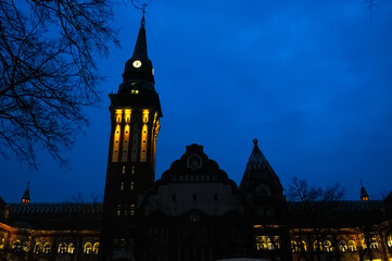 Naklejka premium Illuminated Gradska Kuca city hall in Subotica, Serbia, featuring a clock tower, ornate spires and glowing windows beneath a deep blue evening sky