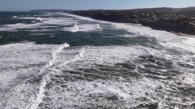 Dynamic drone footage flying forward at a low altitude directly over the rolling white water and crashing waves of the Atlantic Ocean at Praia dos Coxos, Portugal.