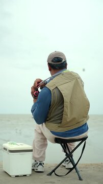 Happy senior man sitting by the sea and fishing with a rod after retirement. Active aging, healthy longevity, outdoor leisure, fulfilling retirement. Life insurance and financial security concept.
