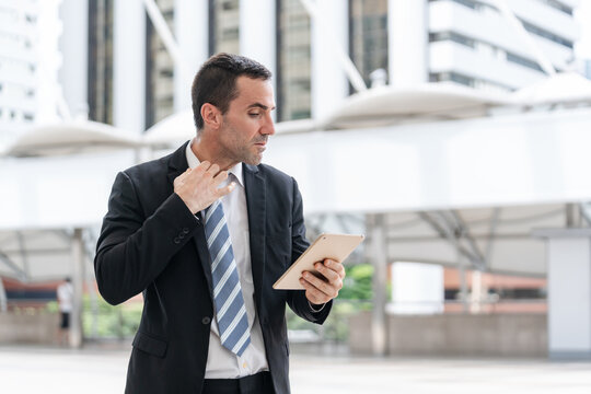 anxious businessman pulling his collar and tie while checking bad news on a digital tablet outdoors office building