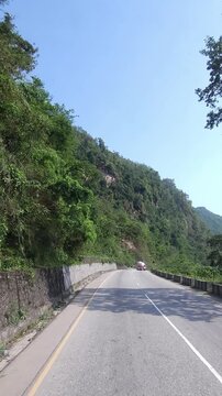 Vertical travel footage showing a winding mountain road passing through dense green forested hills in Nepal. Narrow roadway beside a concrete roadside barrier.