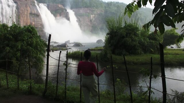 Indian woman admiring the majestic Athirappilly Waterfalls during the monsoon season in Kerala, India, surrounded by lush tropical rainforest and the powerful flow of the Chalakudy River.