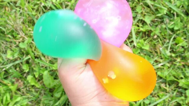 ASMR close-up of two hands holding and squeezing teal, pink and orange water balloons on green grass, pressing the squishy textures together in a slow satisfying motion.